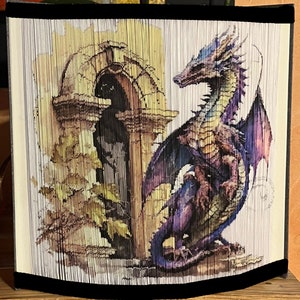 Puede incluir: Una obra de arte de plegado de libros que representa un dragón morado y azul con las alas extendidas. El dragón está de pie frente a un arco de piedra con una ventana. La obra de arte está enmarcada con un borde de terciopelo negro.
