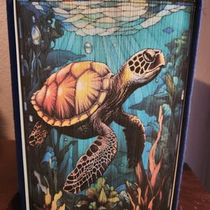 May include: A decorative book with a sea turtle design. The turtle is depicted swimming underwater with a colorful shell and patterned flippers. The background features aquatic plants and bubbles, with a blue and teal color scheme.