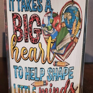 May include: A folded book art piece with a heart shape made of school supplies and the text "It takes a big heart to help shape little minds".
