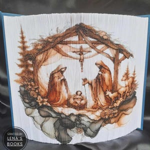 May include: A book sculpture of the Nativity scene, featuring Mary, Joseph, and baby Jesus in a stable, with a cross above. The pages are folded and cut to create the image. The text "Crafted by Lena's Books" is visible on the side of the book.