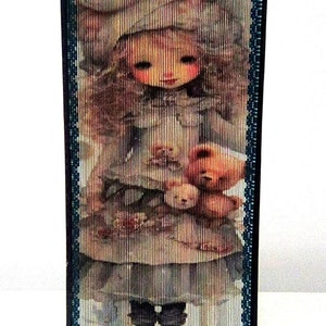 May include: A woven wall hanging featuring a colourful image of a girl holding two teddy bears. The girl is wearing a white hat and a blue dress. The background is a light blue and white.