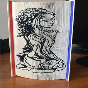 May include: A book art sculpture of a woman's face with a wreath of leaves in her hair. The sculpture is made from folded pages of a book and is painted black. The sculpture is standing on a wooden surface.