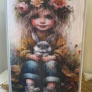 May include: A girl with a flower crown holds a white rabbit in her arms. She is wearing a yellow jacket, blue jeans, and sneakers. The background is a field of flowers.