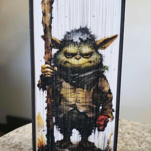 May include: A unique book art piece featuring a green goblin-like character holding a wooden staff and a red object. The artwork is created by meticulously folding the pages of a book, showcasing a detailed illustration with a black border.