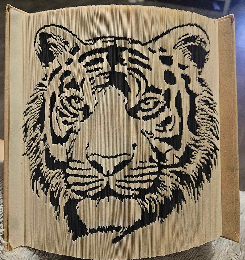 K&ouml;nnte beinhalten: Ein schwarzer und wei&szlig;er Papiertigerkopf, gefaltet aus den Seiten eines Buches. Das Gesicht des Tigers ist mit Streifen und Schnurrhaaren detailliert.