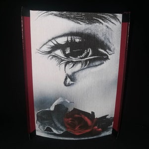May include: A decorative box featuring a black and white illustration of a crying eye and a red rose. The box has a red border and a black background. The artwork is detailed and appears to be a print.