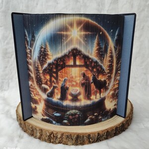 May include: A 3D paper art nativity scene with a star shining above a stable. The scene is surrounded by a glowing, blue, circular frame. The stable is made of wood and has a thatched roof. The figures inside the stable are a mother, father, and baby. There is a donkey and a sheep outside the stable.