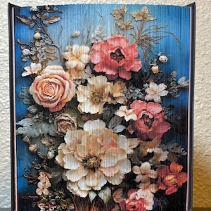 Puede incluir: Una escultura de libro plegado que presenta un vibrante arreglo floral. Las páginas están dobladas con precisión para revelar una imagen colorida de rosas, peonías y varias flores en tonos rosa, crema y naranja sobre un fondo azul intenso.