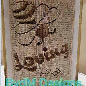 May include: A book art sculpture of a bee with the word "loving" spelled out in the pages. The sculpture is made from a book with the pages folded and cut to create the design. The sculpture is on a white surface with the words "BvdM Designs" in blue text at the bottom.