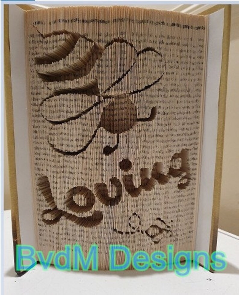 May include: A book art sculpture of a bee with the word "loving" spelled out in the pages. The sculpture is made from a book with the pages folded and cut to create the design. The sculpture is on a white surface with the words "BvdM Designs" in blue text at the bottom.