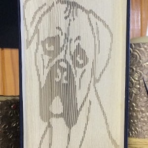 May include: A book sculpture featuring a dog's face, created by precisely folding the pages. The dog's outline is visible against the pages, with the text "vdM Design" at the bottom. The book has a dark blue spine.