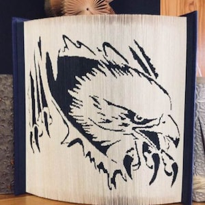 Puede incluir: Un diseño de águila en blanco y negro creado con páginas dobladas de un libro. El águila se muestra con las alas extendidas y las garras extendidas. El libro está encuadernado en tela azul.