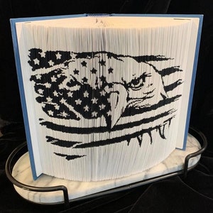 Può includere: Un libro bianco con una copertina blu è piegato in una scultura tridimensionale di un'aquila con una bandiera americana dietro. La scultura è esposta su una base di marmo con una cornice in metallo nero.