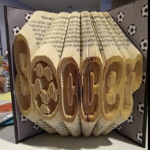 May include: A book folded into the shape of the word "Soccer" with a soccer ball design. The book is displayed on a stand with a black and white patterned fabric.