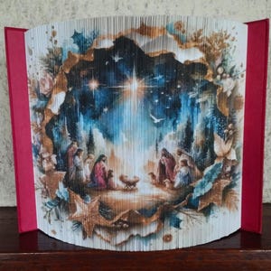 May include: A folded book art piece depicting the nativity scene with a bright star shining down on the manger. The scene is surrounded by a dark blue sky with stars and a golden halo. The book is folded in a way that creates a 3D effect.