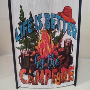 Pode incluir: Um livro branco com uma lombada azul, dobrado em uma escultura 3D. O livro apresenta uma ilustração colorida de uma fogueira, árvores e um chapéu vermelho. O texto "Life is better by the campfire" está impresso no livro.
