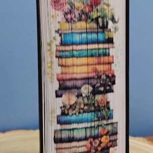 Può includere: Un'opera d'arte unica a forma di libro con una pila di libri con un design floreale. Le pagine sono piegate per creare l'immagine di fiori colorati in cima a una pila di libri. Il libro ha un dorso nero e viene visualizzato in posizione verticale.