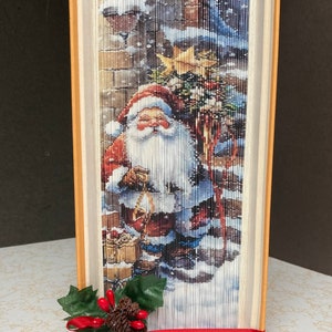 Könnte beinhalten: Eine Buchkunstskulptur des Weihnachtsmanns in einer Schneelandschaft. Das Buch ist zu einer dreidimensionalen Form gefaltet und mit einem roten Band und einem grünen Stechpalmenzweig verziert.