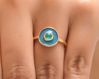 May include: Gold ring with a blue and green enamel design. The ring features a round center with a green stone surrounded by a blue enamel circle.