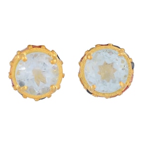 Op de afbeelding: Een paar ronde oorstekers. Elke oorbel heeft een lichtblauwe edelsteen in een goudkleurige zetting. De zetting heeft een decoratieve rand met kleine gouden accenten en gekleurde stenen. De oorbellen worden tegen een witte achtergrond getoond.