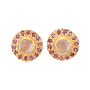 Puede incluir: Un par de pendientes de botón dorados. Cada pendiente presenta una piedra central de color claro rodeada por un anillo de pequeñas piedras preciosas rosas. Los pendientes tienen un diseño circular.