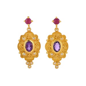 May include: A pair of gold-colored earrings with an ornate design. Each earring features a central oval amethyst stone, surrounded by small pearls and a ruby at the top. The earrings have a vintage, regal aesthetic.