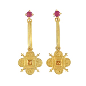May include: Gold hoop earrings with a dangling square charm featuring a small orange gemstone. The charm is surrounded by intricate gold detailing and the earrings are topped with a small red gemstone.
