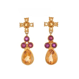 May include: Gold earrings with a cross design and teardrop-shaped orange gemstones. The cross is adorned with smaller, clear gemstones. The teardrop gemstones are accented with four smaller, purple gemstones.