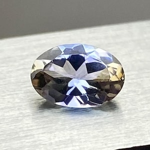 May include: An oval-shaped gemstone with multiple facets, exhibiting a blend of blue, purple, and golden-brown tones. The gem is presented against a plain backdrop, highlighting its clarity and sparkle. The gemstone's form and colour variations are noticeable.