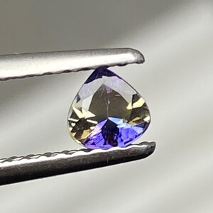 May include: A heart-shaped gemstone, held by tweezers, shows a mix of colours. The gem has a deep blue base, fading to golden-brown at the top. The facets reflect light, creating a sparkling effect. A beautiful gemstone.