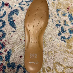May include: A tan-colored Clarks Active Air insole with small ventilation holes. The insole is designed to fit inside a shoe and has the brand name "Clarks" printed on it. The insole is made of a soft material.
