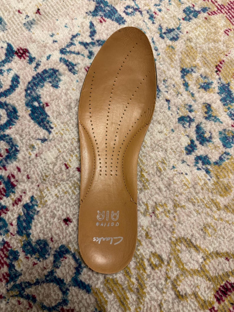 May include: A tan-colored Clarks Active Air insole with small ventilation holes. The insole is designed to fit inside a shoe and has the brand name "Clarks" printed on it. The insole is made of a soft material.