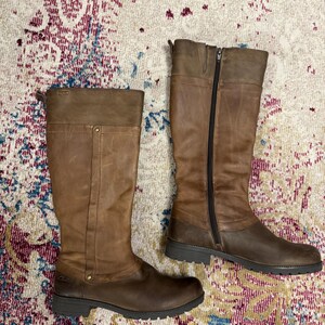May include: A pair of brown leather knee-high boots. One boot is zipped up, and the other is unzipped. The boots have a dark brown sole and a lighter brown upper. The boots are on a patterned rug.