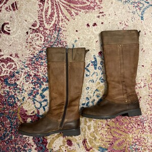May include: A pair of brown leather knee-high boots. One boot has a full-length zipper. The boots have a two-tone design, with a lighter brown on the upper shaft and a darker brown on the lower portion and sole. The boots are on a patterned rug.