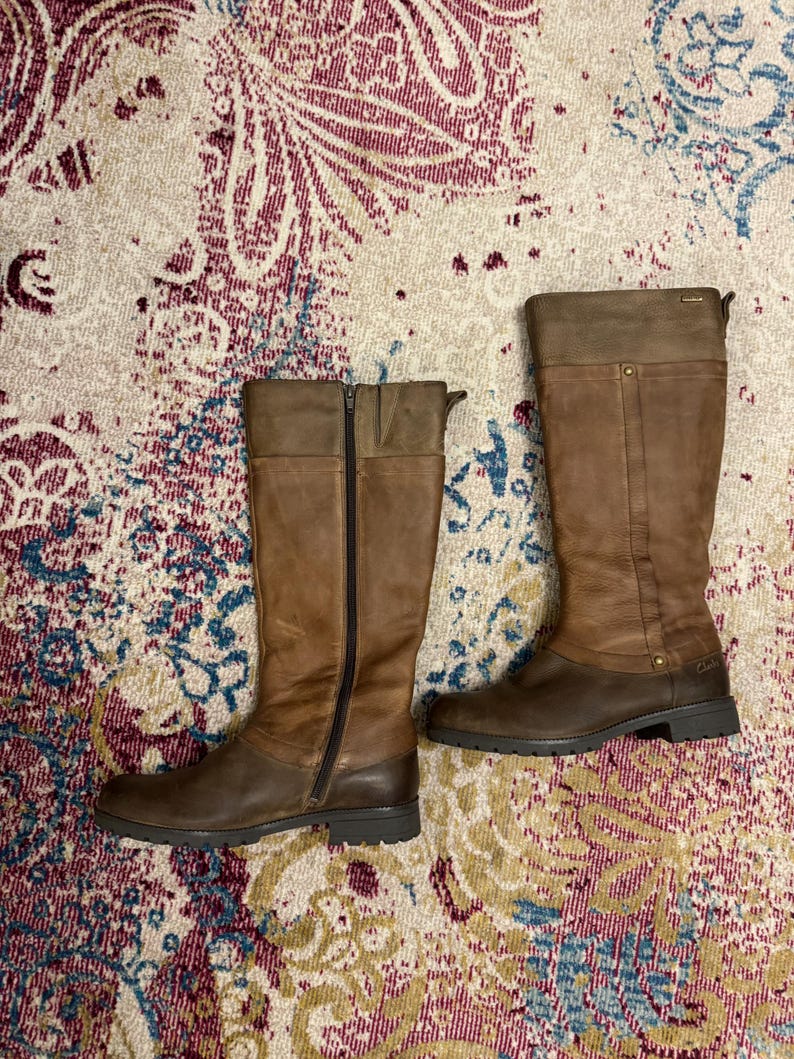 May include: A pair of brown leather knee-high boots. One boot has a full-length zipper. The boots have a two-tone design, with a lighter brown on the upper shaft and a darker brown on the lower portion and sole. The boots are on a patterned rug.
