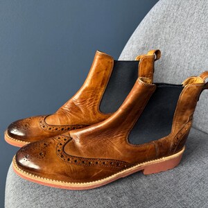 May include: A pair of tan leather Chelsea boots featuring brogue detailing on the toe cap and sides. The boots have black elastic side panels, pull tabs at the heels, and a flat, reddish-brown sole with a low heel for a classic look.