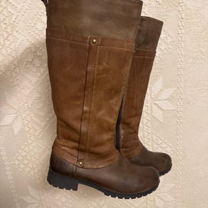 May include: A pair of brown leather knee-high boots. The boots have a two-tone design, with a darker brown at the base and a lighter brown at the top. They feature a side zipper and a small heel.
