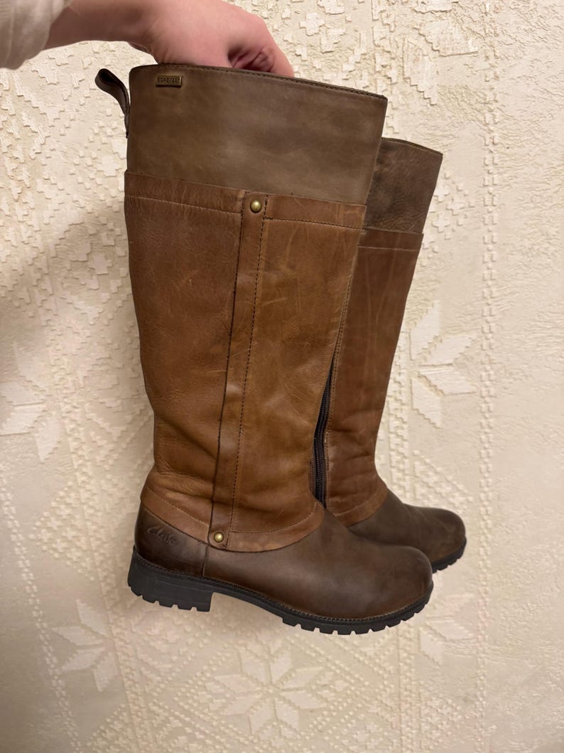 May include: A pair of brown leather knee-high boots. The boots have a two-tone design, with a darker brown at the base and a lighter brown at the top. They feature a side zipper and a small heel.