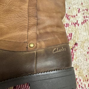 May include: Close-up of a brown leather boot with a dark brown sole. The boot has a textured appearance with visible stitching and a gold-colored rivet. The brand name "Clarks" is subtly embossed on the side.