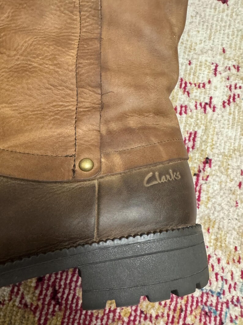 May include: Close-up of a brown leather boot with a dark brown sole. The boot has a textured appearance with visible stitching and a gold-colored rivet. The brand name "Clarks" is subtly embossed on the side.