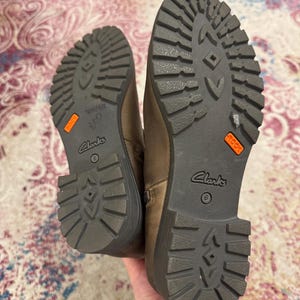 May include: Pair of brown leather Clarks boots, size 6, with dark gray soles. The soles have a rugged tread pattern and an orange "ROCK" label. The boots are held up by a hand.