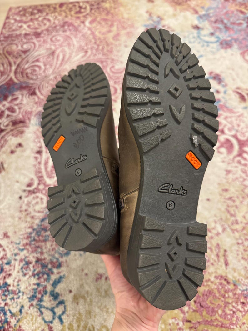 May include: Pair of brown leather Clarks boots, size 6, with dark gray soles. The soles have a rugged tread pattern and an orange "ROCK" label. The boots are held up by a hand.