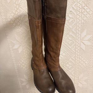 May include: A pair of brown leather knee-high boots. The boots have a two-tone design, with a lighter brown on the upper shaft and a darker brown on the lower part. They feature a zipper closure and a rounded toe.