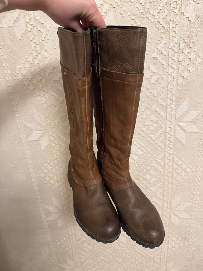 May include: A pair of brown leather knee-high boots. The boots have a two-tone design, with a lighter brown on the upper shaft and a darker brown on the lower part. They feature a zipper closure and a rounded toe.