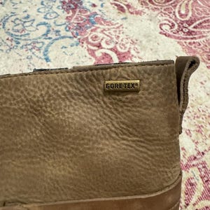 May include: Close-up of a brown leather boot with a textured surface. The boot features a metal tag that reads "GORE-TEX". The boot has a stitched design and a brass-colored button.