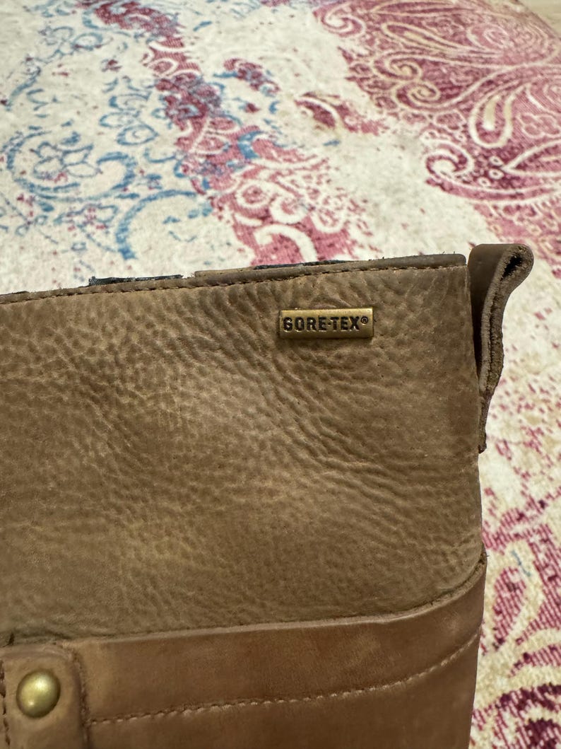 May include: Close-up of a brown leather boot with a textured surface. The boot features a metal tag that reads "GORE-TEX". The boot has a stitched design and a brass-colored button.