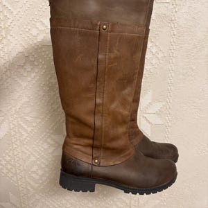 May include: A pair of brown leather knee-high boots. The boots have a two-tone design, with a darker brown lower section and a lighter brown upper section. The boots have a round toe and a rubber sole.