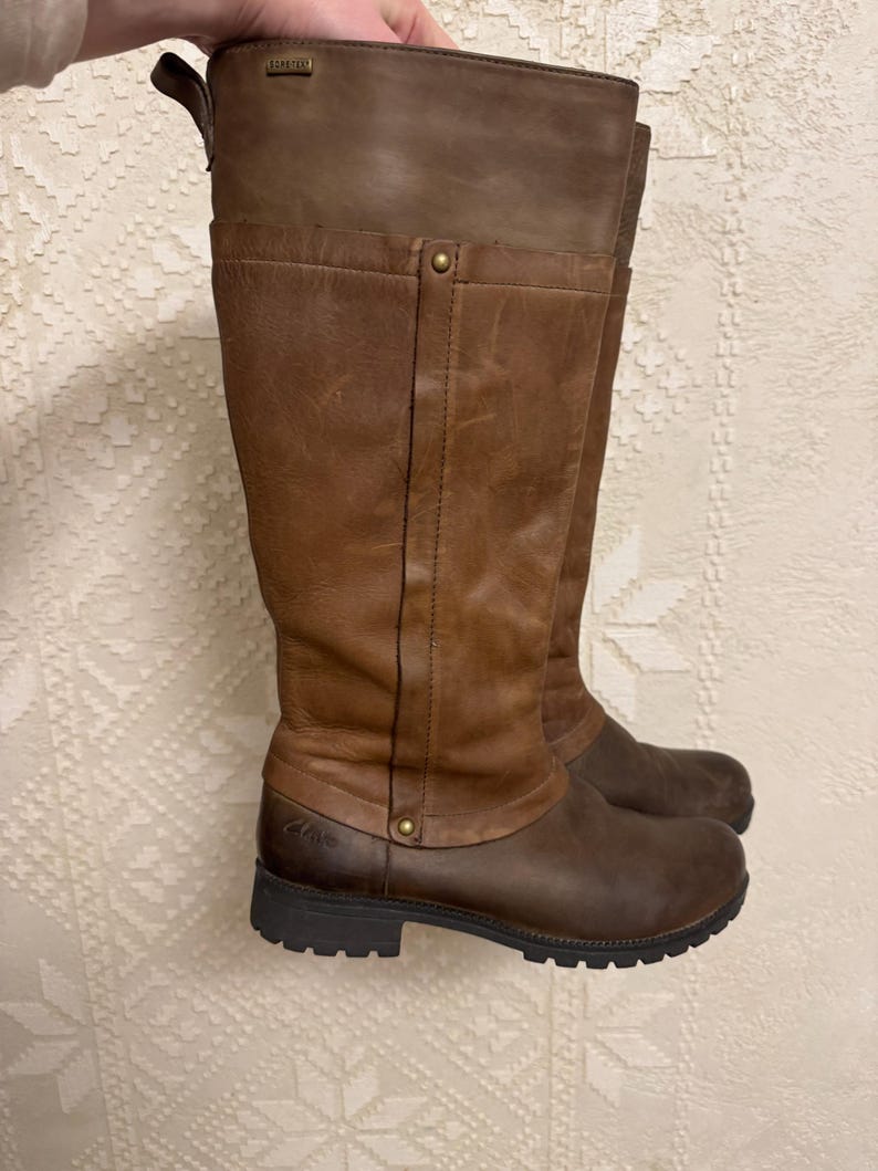 May include: A pair of brown leather knee-high boots. The boots have a two-tone design, with a darker brown lower section and a lighter brown upper section. The boots have a round toe and a rubber sole.