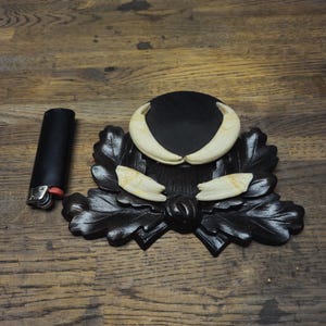 May include: A black carved wooden decorative tray with a leaf design, holding a black circular object with two large, curved, off-white tusks. Two smaller tusks are also present. A black lighter is on the left.