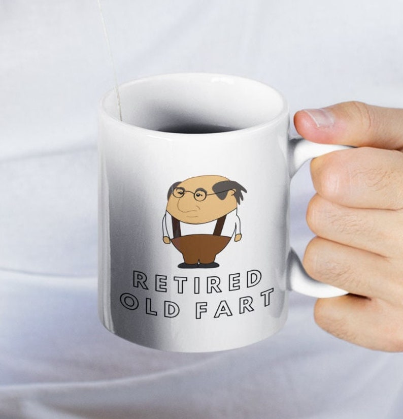 K&ouml;nnte beinhalten: Wei&szlig;e Keramik-Tasse mit einer Cartoon-Illustration eines Mannes mit Brille und dem Text "RETIRED OLD FART". Die Illustration zeigt den Mann in einem wei&szlig;en Hemd, brauner Hose und Hosentr&auml;gern. Die Tasse hat einen wei&szlig;en Henkel.